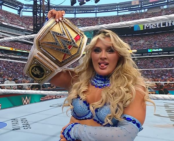 A female wrestler, Tiffany Stratton, holds up the WWE Championship belt triumphantly at WWE Summerslam. She has long blonde hair and is wearing a sparkling blue outfit.