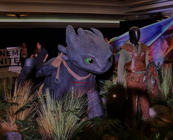 A large, animated dragon character with bright green eyes stands among foliage, accompanied by a mannequin dressed in medieval clothing. The scene suggests a fantasy theme, possibly related to a popular animated series or film.