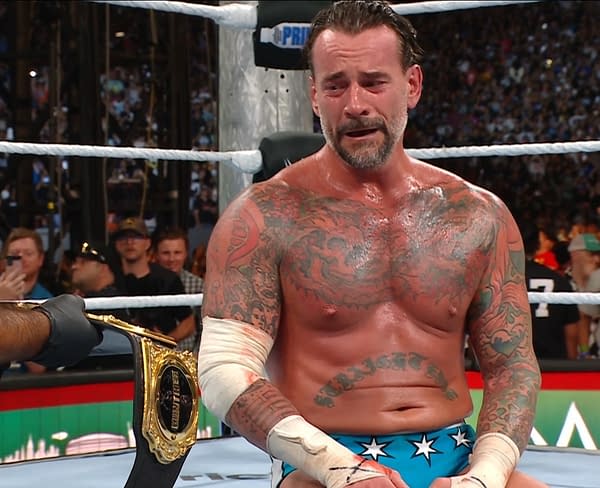 A muscular wrestler with extensive tattoos, CM Punk, sits inside a wrestling ring, visibly emotional after a match, with a championship belt nearby. The crowd in the background reacts to the event.