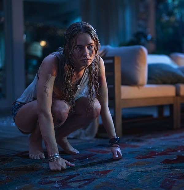 Johnny Sequoyah as "Lucy" crouches on a patterned rug in a dimly lit room, showcasing a tense expression and disheveled hair, reflecting a dramatic moment from the film "Primate".