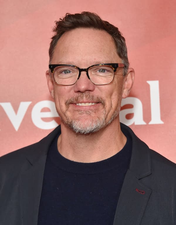 Matthew Lillard smiles at the camera, wearing glasses and a blazer, at a red carpet event, with a soft-focus background in shades of pink and orange.