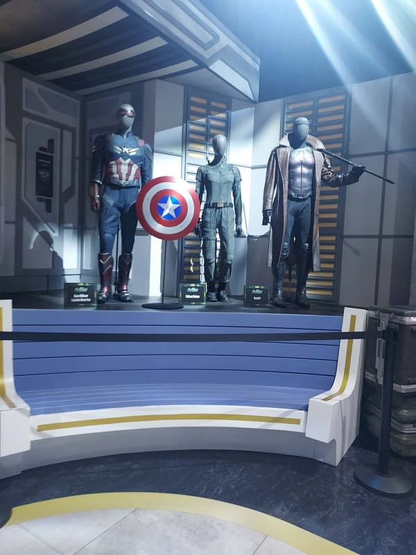 Three Avengers: Doomsday costumes on display at SDCC, including Captain America with a shield and two other superhero suits on a futuristic stage.