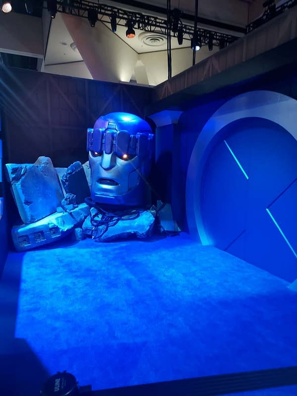 A blue-lit Comic-Con display of a giant Sentinel head emerging from rubble, with a futuristic circular backdrop.
