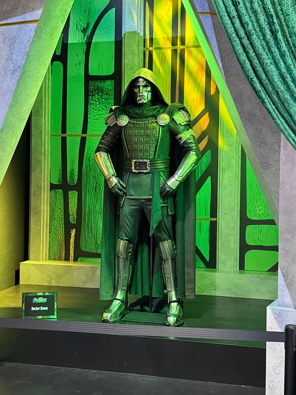 A Doctor Doom display figure stands in a green-lit Avengers: Doomsday SDCC exhibit, wearing a hooded mask and metallic armor.