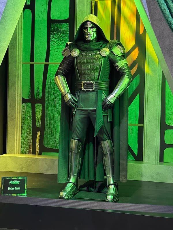 Doctor Doom costume display at SDCC, with a hooded green-and-silver armored figure standing on a stage. A small sign at the base identifies the Avengers: Doomsday exhibit.