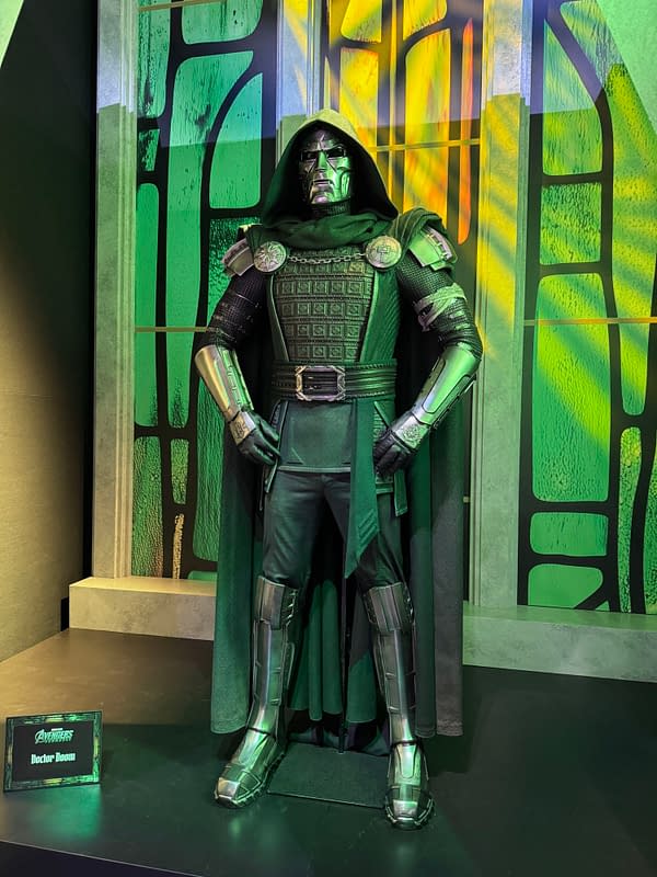 Doctor Doom costume on display at SDCC, with a hooded metal mask, dark green armor, and a cape against a glowing green-and-yellow backdrop.