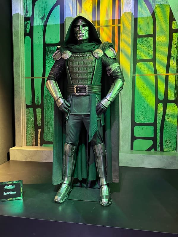 A Doctor Doom costume display at SDCC, featuring the armored Marvel villain in a green hooded cloak and silver mask. A small sign reads