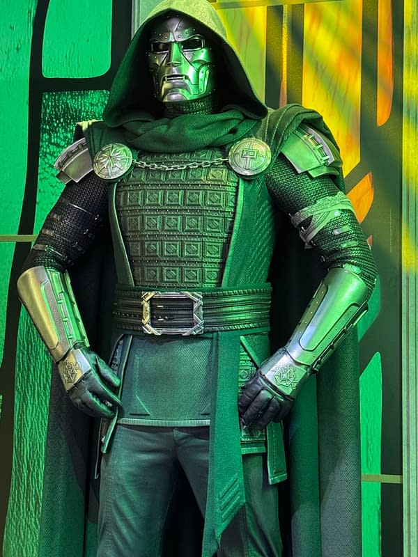 A life-size Doctor Doom costume with a hooded cloak, silver mask, and green-lit armor displayed at San Diego Comic-Con. The figure stands against a dramatic yellow and green backdrop.