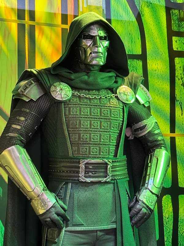 A person in a Doctor Doom costume stands against a green-lit Avengers: Doomsday display at SDCC, wearing a hooded cloak, metal mask, and armored suit.