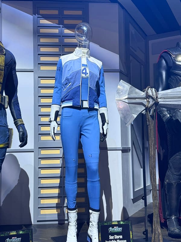 Fantastic Four costume display featuring Sue Storm's blue-and-white suit from Avengers: Doomsday at SDCC, shown beside other character costumes and a large Sentinel prop.