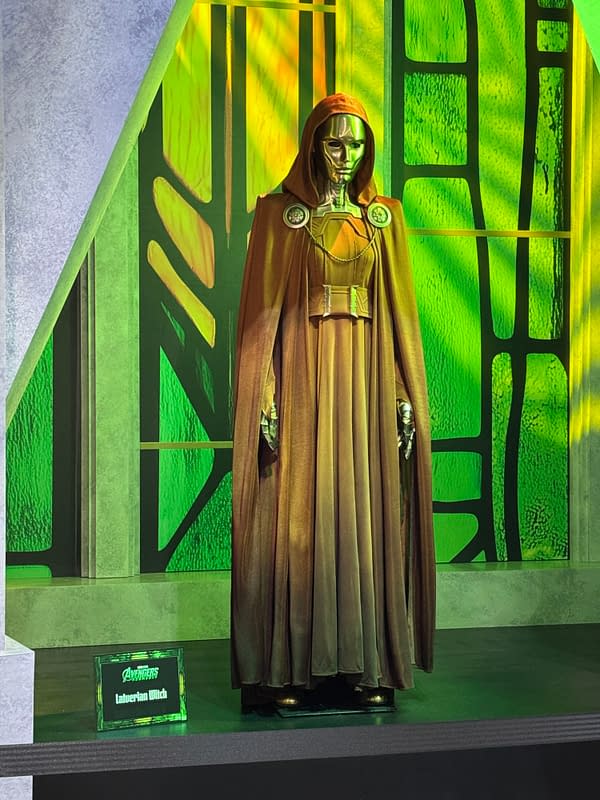 A hooded Latverian Witch costume from Avengers: Doomsday stands on display at SDCC, featuring a gold mask and brown cloak against a glowing green backdrop.