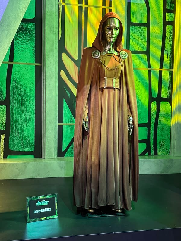 A display of a Latverian Witch costume from Avengers: Doomsday at SDCC, featuring a hooded metallic mask and flowing brown cloak against a green-lit backdrop.