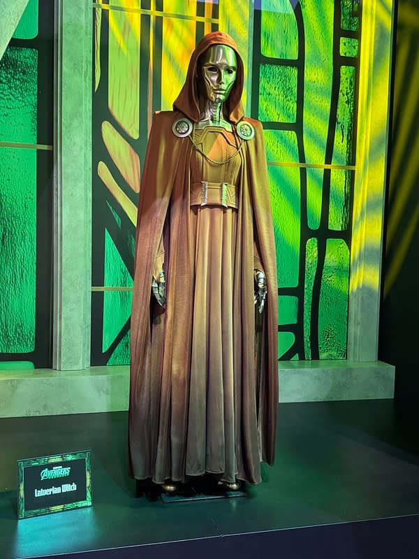 A Latverian Witch costume from Avengers: Doomsday stands on display at SDCC, featuring a metallic mask, hooded brown cloak, and green-lit backdrop.