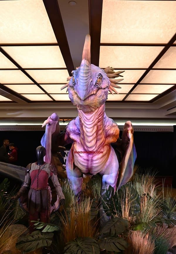 A large, colorful dragon-like creature stands prominently in a convention setting, surrounded by greenery. A mannequin dressed in medieval armor is positioned nearby, adding a fantasy theme to the scene.