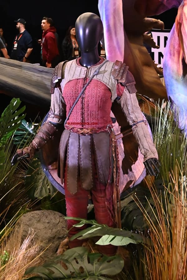 A detailed costume display features a mannequin wearing a textured, red outfit with leather armor pieces and accessories. The background includes lush greenery, enhancing the visual impact of the costume's design.