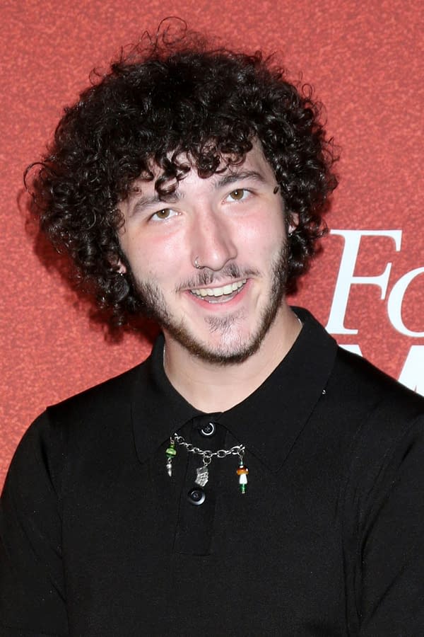 Franklin Jonas, a young man with curly hair and a beard, smiles at the camera, wearing a black polo shirt adorned with a necklace of colored charms. The background features a vibrant red surface, suggesting a festive atmosphere.