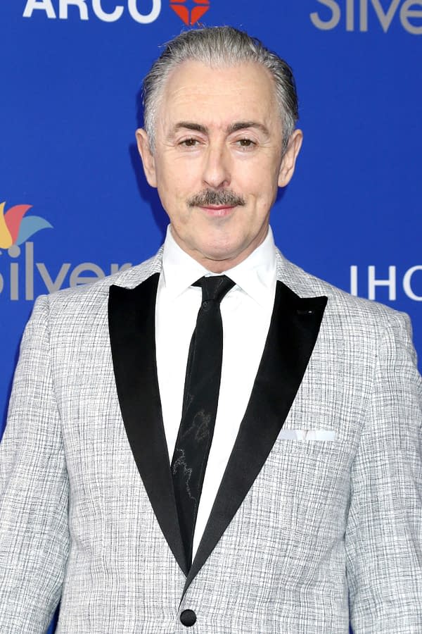 A man with gray hair and a mustache smiles while wearing a stylish gray suit with a black lapel and a white shirt. The background features logos, creating a celebratory atmosphere.