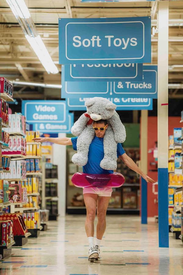A man humorously dressed in a blue shirt and pink shorts, wearing sunglasses, struts down a store aisle while balancing a large stuffed animal on his shoulders and a pink inflatable ring around his waist. The store's signage prominently displays sections for soft toys and video games.