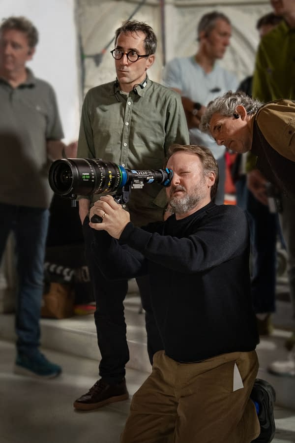 On the set of 'Wake Up Dead Man: A Knives Out Mystery,' Writer/Director Rian Johnson is kneeling and adjusting a camera lens, while Director of Photography Steve Yedlin watches intently. The background features crew members and filming equipment.