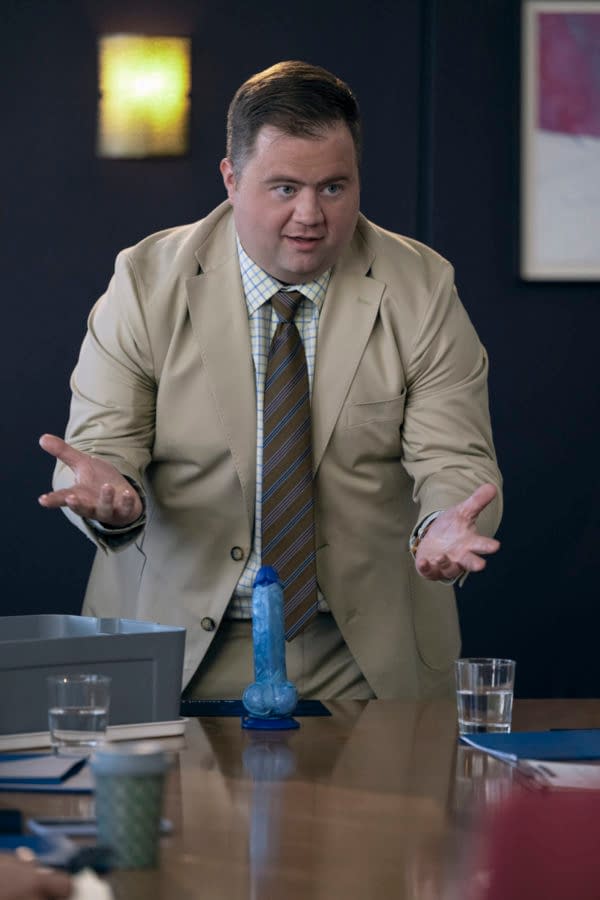 A man wearing a beige suit and a checkered shirt gestures with his hands while presenting a humorous blue object in a meeting room setting. The image captures a comedic moment from the show 'Balls Up'.
