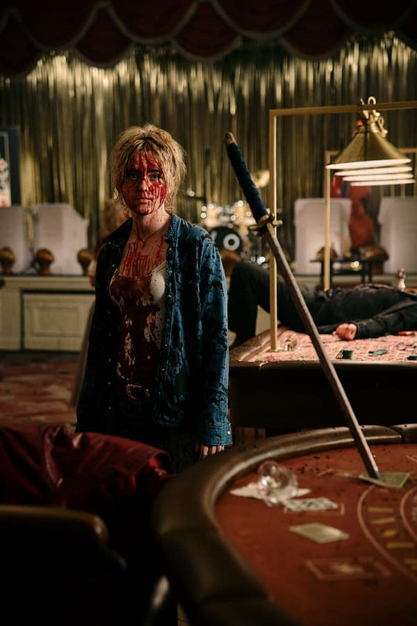 Kathryn Newton in a scene from 'Ready or Not 2,' appears disheveled and bloody, standing in a dimly lit, chaotic environment with a sword nearby and a casino table scattered with cards and money.