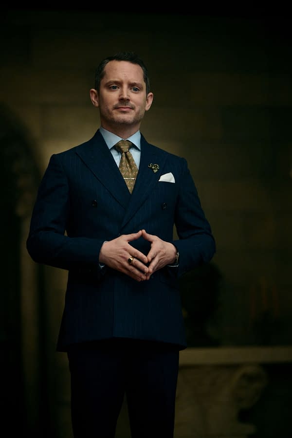 Elijah Wood stands confidently in a dark suit with a gold tie, hands clasped together in front of him, as he appears in the film READY OR NOT 2: HERE I COME.