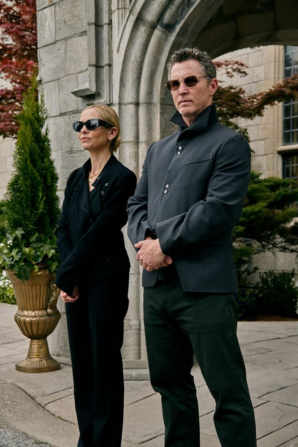 Sarah Michelle Gellar and Shawn Hatosy stand together in stylish outfits, both wearing sunglasses, outside a stone archway in a garden setting, promoting the film 'READY OR NOT 2: HERE I COME'.