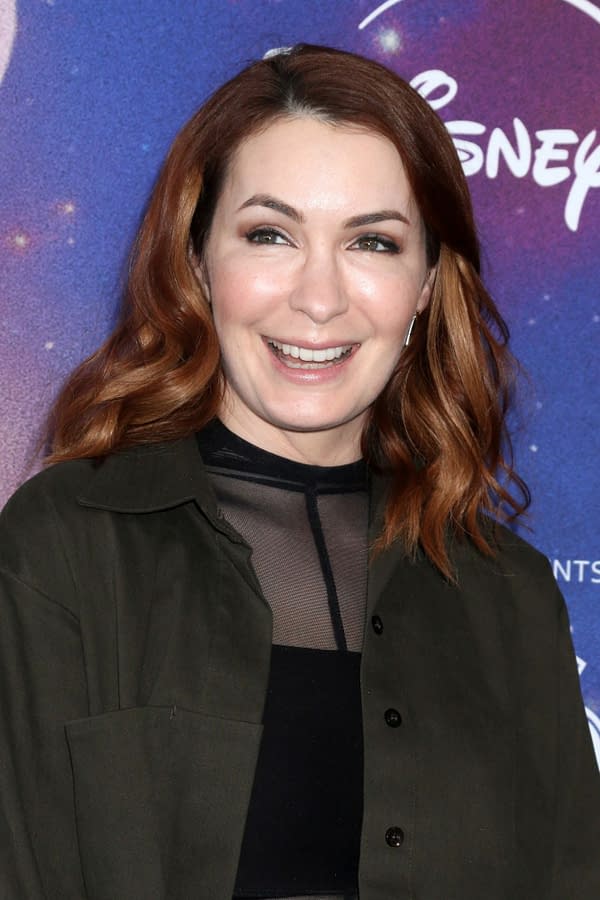 Felicia Day appears on the red carpet, smiling widely for the camera. She has long, wavy hair and is wearing a dark jacket over a black top.