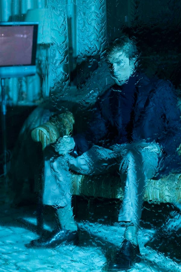 Blue-tinted, rain-streaked view of a person standing indoors beside a striped chair, seen through wet glass.