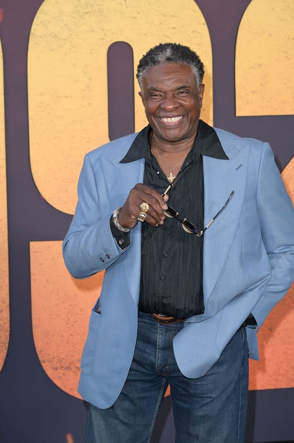 Keith David smiles at a Los Angeles premiere, wearing a light blue blazer over a black shirt and jeans while holding his glasses.
