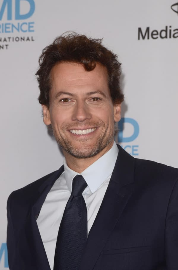 A man with curly hair and a beard smiles at the camera while wearing a dark suit and a light blue shirt, set against a backdrop with logos for IMDb and Media. This image is associated with discussions about Ioan Gruffudd's involvement in the original 'Fantastic Four' trilogy.