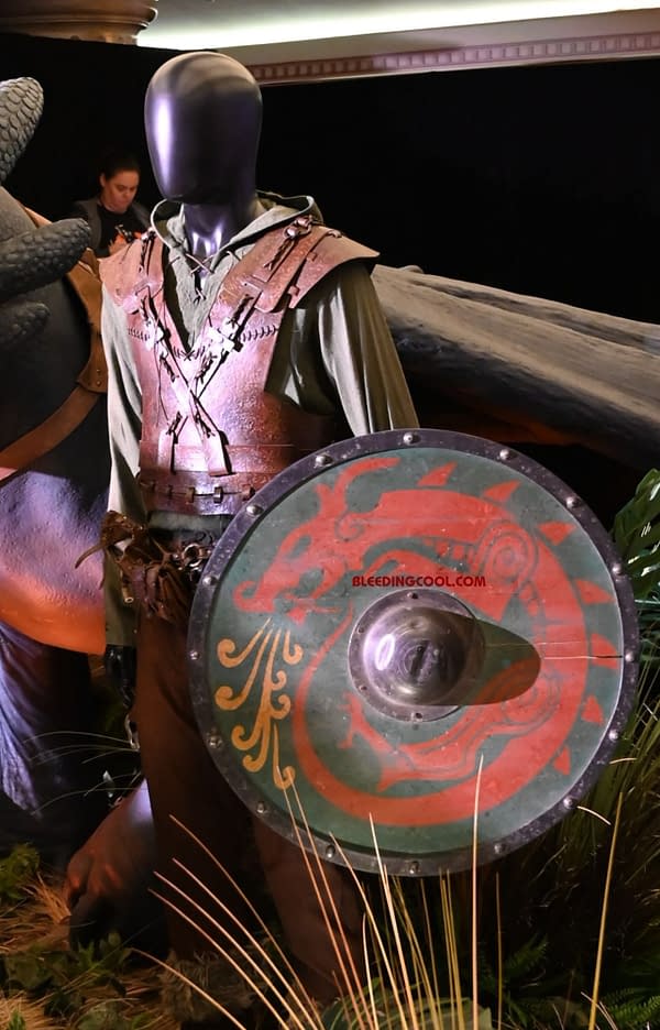 A mannequin dressed in a medieval costume featuring a green hooded tunic and protective leather armor, holding a painted shield with a dragon design. A large dragon figure is partially visible in the background.